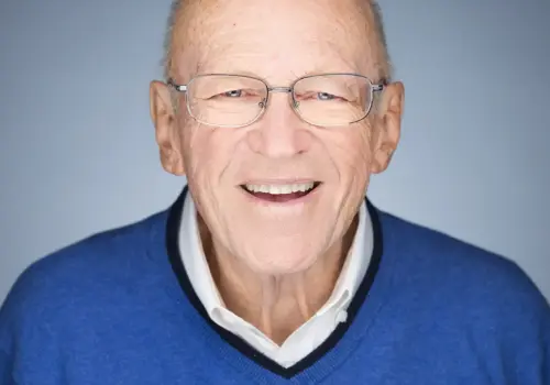 Ken Blanchard, ein älterer Mann mit Brille und Glatze, lächelt warmherzig. Er trägt einen blauen Pullover über einem weißen Hemd mit Kragen und steht vor einem schlichten hellblauen Hintergrund.