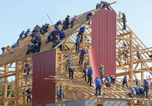 Dutzende von Menschen arbeiten gemeinsam am Holzgerüst einer großen Scheune und montieren unter strahlend blauem Himmel rote Metallpaneele auf dem Dach und an den Wänden, wobei die psychologische Sicherheit am Arbeitsplatz durch die effiziente Zusammenarbeit gefördert wird.