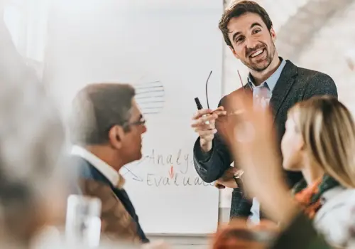 A man in a suit smiles and gestures while presenting learning myths in continuing education to colleagues in a bright office. He stands before a flipchart with graphs and notes, engaging with seated team members.