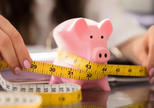 Eine Person wickelt ein gelbes Maßband um ein rosa Sparschwein auf einem Schreibtisch und symbolisiert damit Budgetierung, finanzielle Ersparnisse und die Bedeutung der Weiterbildung in Krisenzeiten.