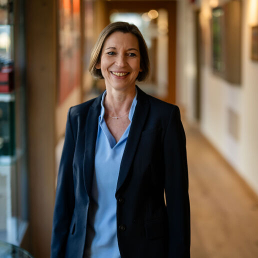 Eine Frau mit kurzen braunen Haaren, einer blauen Bluse und einem dunklen Blazer steht lächelnd in einem gut beleuchteten Flur mit Kunstwerken und Fotos an den Wänden.