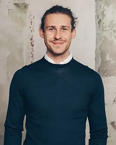 Vincent Aretz. ein Mann mit langen, zurückgebundenen Haaren und einem kurzen Bart lächelt in die Kamera. Er trägt einen dunklen Pullover über einem weißen Kragenhemd und steht vor einer strukturierten, verwitterten Wand.