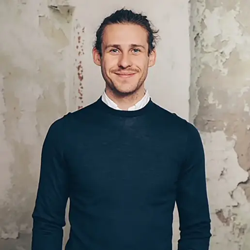 Vincent Aretz. ein Mann mit langen, zurückgebundenen Haaren und einem kurzen Bart lächelt in die Kamera. Er trägt einen dunklen Pullover über einem weißen Kragenhemd und steht vor einer strukturierten, verwitterten Wand.