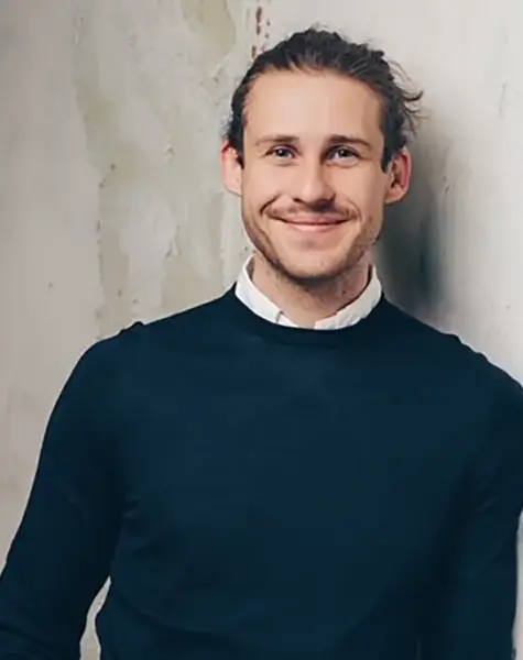 Vincent Aretz, ein Mann mit hellem Bart und langen, nach hinten gebundenen Haaren lehnt lächelnd an einer hellen, strukturierten Wand. Er trägt einen dunklen Pullover über einem weißen Hemd mit Kragen.