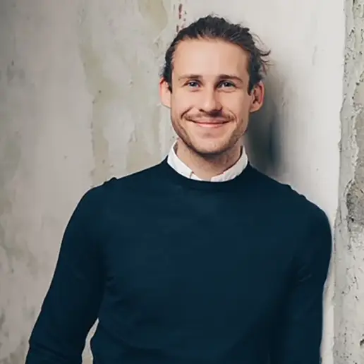 Vincent Aretz, ein Mann mit hellem Bart und langen, nach hinten gebundenen Haaren lehnt lächelnd an einer hellen, strukturierten Wand. Er trägt einen dunklen Pullover über einem weißen Hemd mit Kragen.