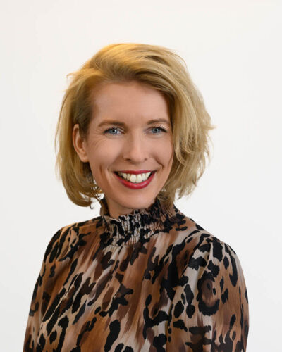 Hanneke Kersting, eine Frau mit blonden, schulterlangen Haaren, lächelt in die Kamera. Sie trägt eine hochgeschlossene Langarmbluse mit braun-schwarzem Leopardenmuster auf schlichtem weißem Grund.