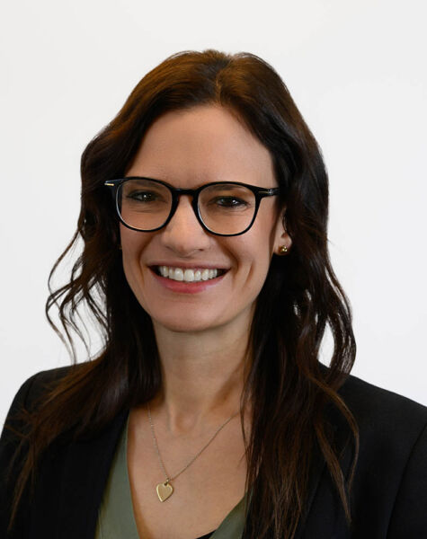 Sonja Herr, eine Frau mit langen braunen Haaren und Brille, trägt einen dunklen Blazer über einem grünen Oberteil und eine herzförmige Halskette und lächelt vor einem schlichten weißen Hintergrund.