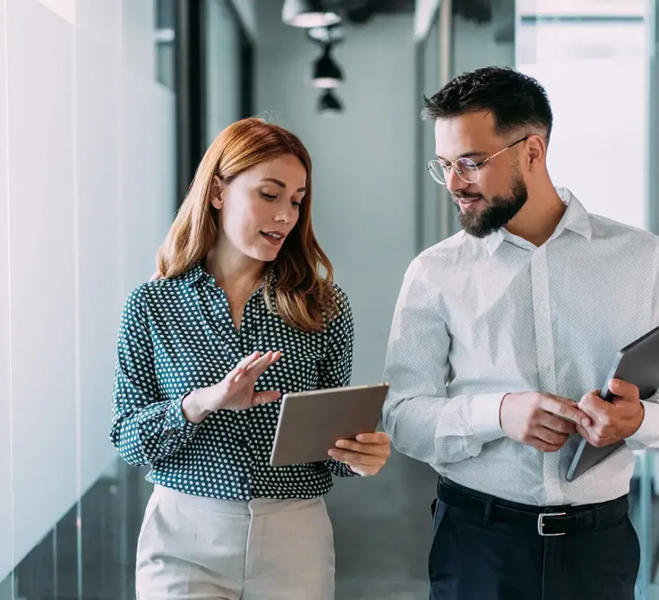 Zwei Geschäftsleute gehen souverän durch einen Büroflur und prüfen Informationen auf einem Tablet. Beide tragen Business-Kleidung. Die Frau deutet auf das Tablet, der Mann hält einen Ordner in der Hand und blickt auf den Bildschirm.