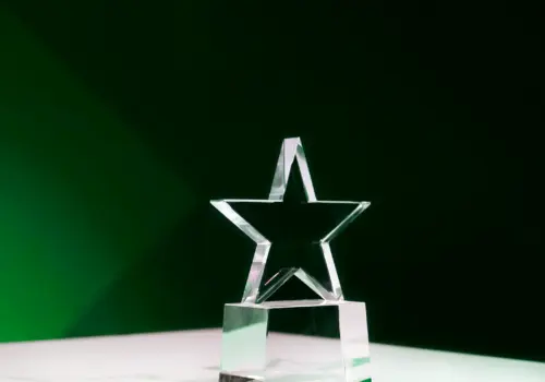 Eine sternförmige Trophäe aus klarem Glas steht auf einer weißen Oberfläche mit dunkelgrünem Hintergrund und wird von sanftem Licht beleuchtet.