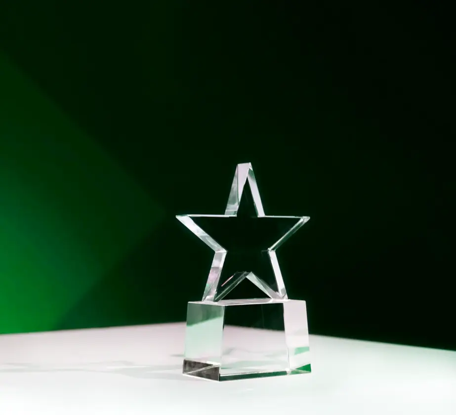 Eine sternförmige Trophäe aus klarem Glas steht auf einer weißen Oberfläche mit dunkelgrünem Hintergrund und wird von sanftem Licht beleuchtet.