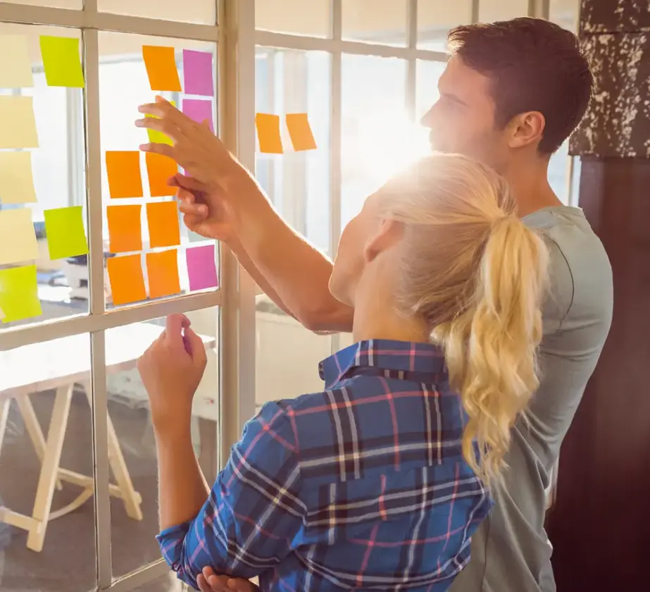Zwei Personen stehen an einem mit bunten Haftnotizen bedeckten Fenster und führen ein Brainstorming durch. Einer zeigt auf die Notizen, der andere schaut nachdenklich zu, während das Sonnenlicht hinter ihnen hereinströmt.