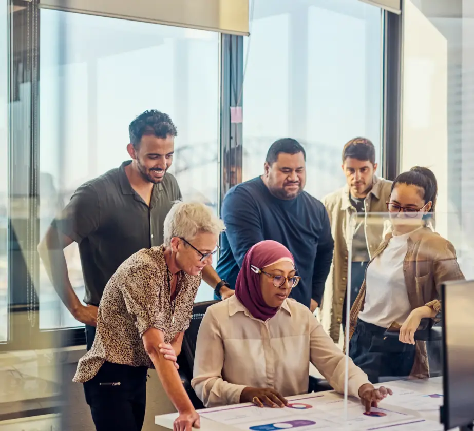 Eine vielfältige Gruppe von fünf Personen arbeitet mit Diagrammen und Dokumenten an einem Schreibtisch zusammen, diskutiert und trainiert Inklusion, während sie in einem modernen Büro mit großen Fenstern auf einen Computerbildschirm blickt.