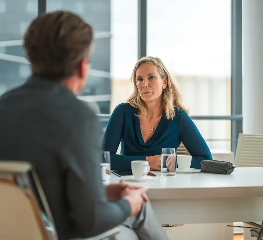 Eine Frau in einer blauen Bluse sitzt an einem Tisch und hört einem Mann ihr gegenüber aufmerksam zu. Sie befinden sich in einem modernen Büro mit großen Fenstern. Auf dem Tisch stehen Tassen, Gläser Wasser und ein Beutel.