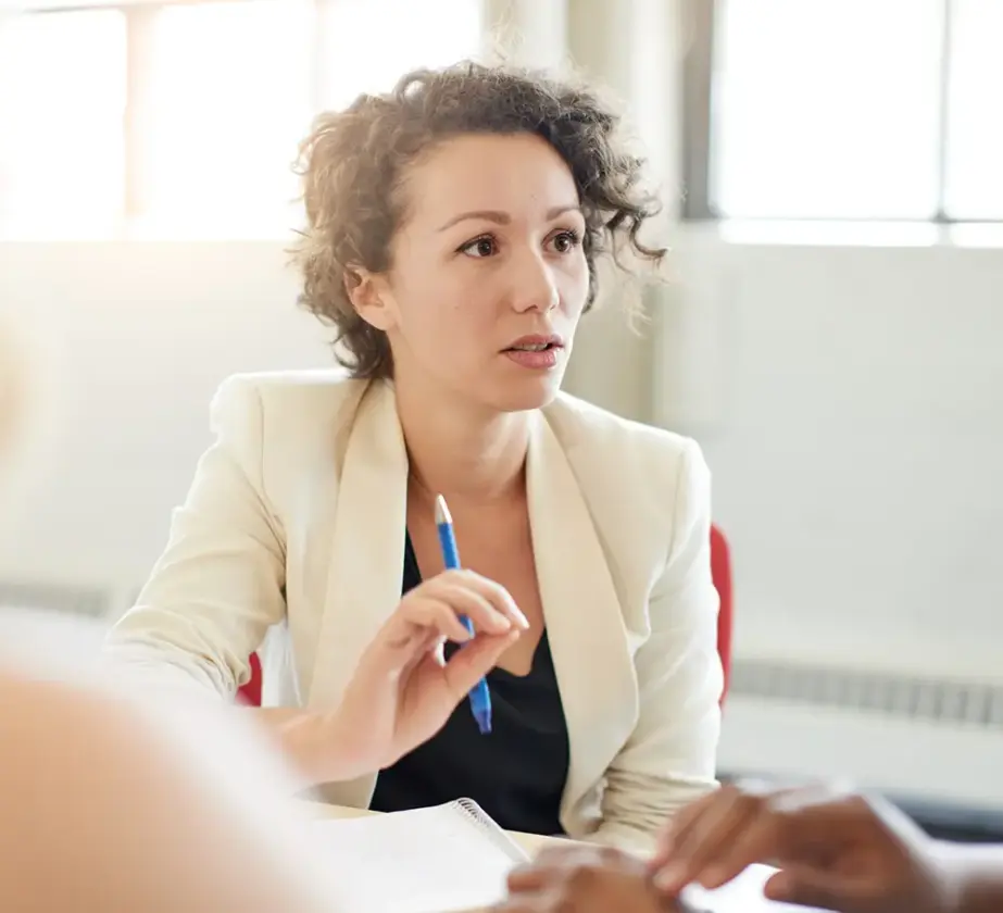 Eine Frau mit lockigem Haar und einem hellen Blazer sitzt mit einem Stift in der Hand an einem Tisch in einem hellen Raum mit großen Fenstern. Sie scheint in eine Diskussion oder Besprechung vertieft zu sein.