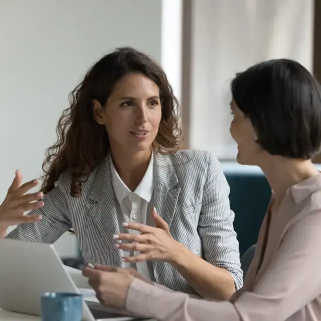 Zwei Frauen sitzen an einem Tisch und diskutieren über Präsentationscoaching. Die eine gestikuliert, die andere hört aufmerksam zu. Vor ihnen im Büro oder Besprechungsraum stehen ein Laptop und eine blaue Tasse.