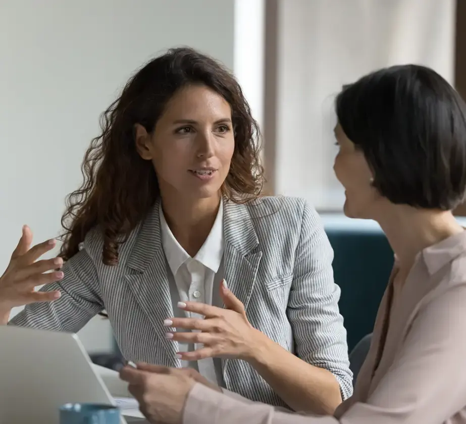 Zwei Frauen sitzen an einem Tisch und diskutieren über Präsentationscoaching. Die eine gestikuliert, die andere hört aufmerksam zu. Vor ihnen im Büro oder Besprechungsraum stehen ein Laptop und eine blaue Tasse.