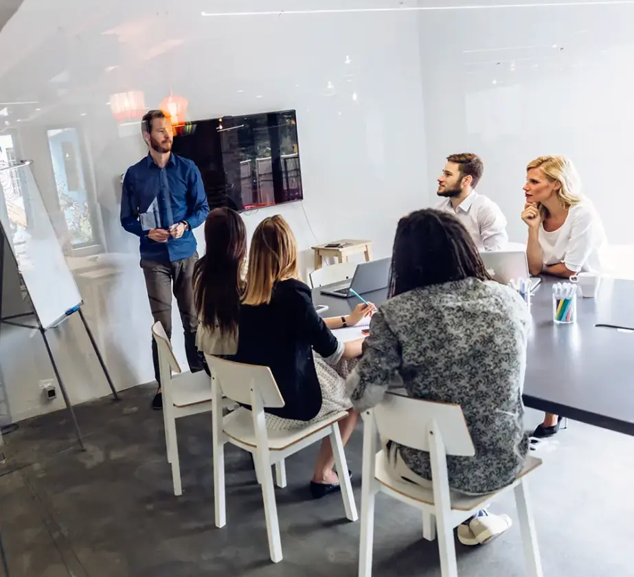 Ein Mann steht an einem Whiteboard und erklärt vier Kollegen, die in einem modernen Büro-Besprechungsraum um einen Konferenztisch sitzen, wie man pyramidenförmig kommuniziert. Hinter ihm ist an der Wand ein großer Monitor angebracht.