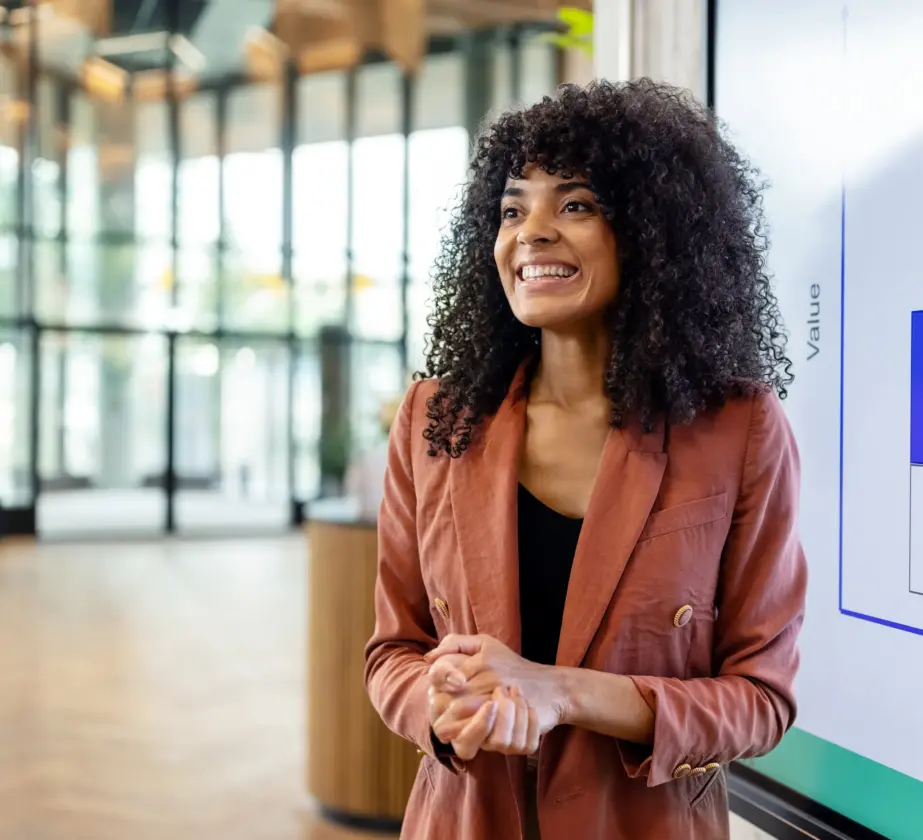 A woman with curly hair, wearing a rust-colored blazer, stands smiling in front of a presentation screen displaying a chart, in a modern, bright office setting.