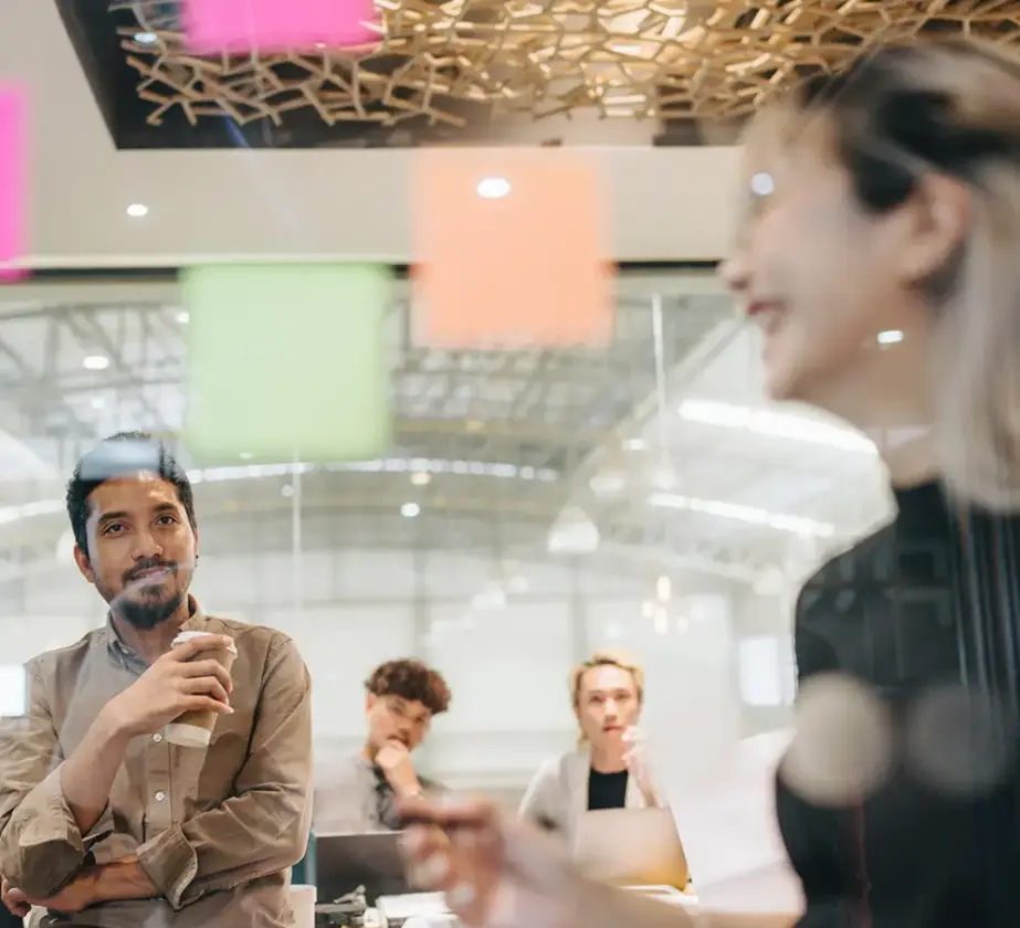 Vier Personen in einem modernen Büro führen ein gemeinsames Brainstorming durch und nutzen Storytelling, um Ideen zu entwickeln. Eine Person steht lächelnd mit einem Stift in der Hand da, während andere sitzen und zuhören. Im Vordergrund kleben bunte Haftnotizen an einer Glaswand.
