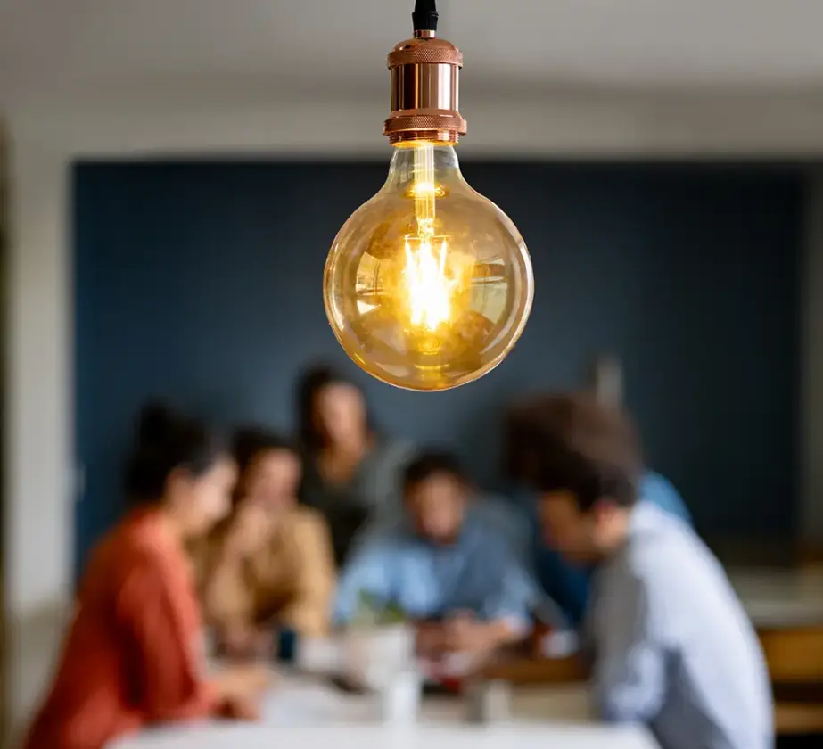 Im Fokus steht eine brennende hängende Glühbirne, im unscharfen Hintergrund sitzt eine Gruppe von vier Personen, die an einem Training zur Teamkommunikation teilnehmen, um einen Tisch herum und unterhält sich.