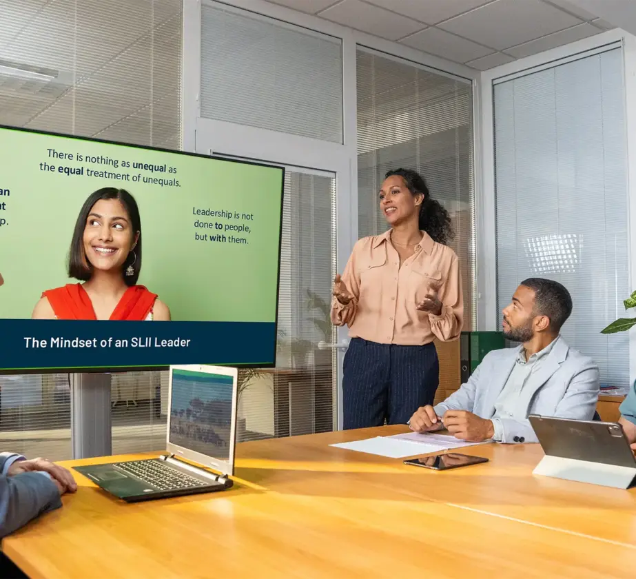 Four colleagues in business attire are having a meeting in a modern conference room. A woman stands and presents information about leadership from a screen, while three others listen and take notes.