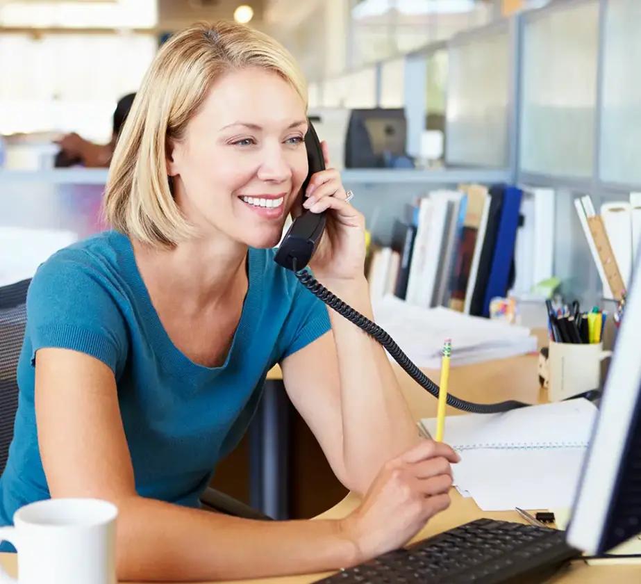 Eine Frau sitzt lächelnd an ihrem Schreibtisch in einem Büro, während sie in ein schnurgebundenes Telefon telefoniert und auf ihren Computermonitor schaut – ein perfekter Eindruck von der Konzentration und Energie, die man oft bei Verkaufstrainings oder Telefonvertriebssitzungen sieht.