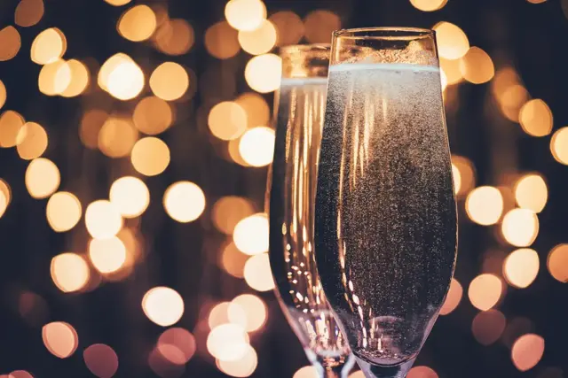Im Fokus stehen zwei mit Sekt gefüllte Champagnergläser, vor einem warmen, goldenen Bokeh-Hintergrund aus verschwommenen Lichtern, der eine festliche Atmosphäre schafft.