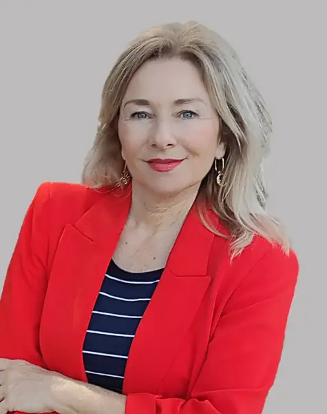 Trainerin Andrea König-Wenskus, Expertin für Mensch & Kommunikation, mit langen blonden Haaren im roten Blazer über schwarz-weißem Streifenshirt, steht mit verschränkten Armen und lächelt vor grauem Hintergrund.