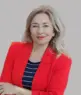 Trainerin Andrea König-Wenskus, Expertin für Mensch & Kommunikation, mit langen blonden Haaren im roten Blazer über schwarz-weißem Streifenshirt, steht mit verschränkten Armen und lächelt vor grauem Hintergrund.