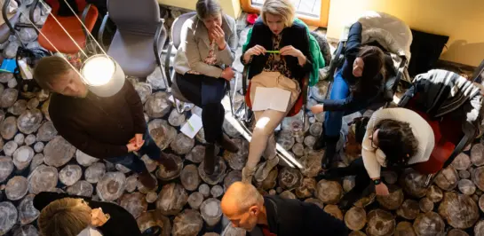 Eine Gruppe von Menschen sitzt und steht in einem gemütlichen Besprechungsraum mit Steinboden (von oben betrachtet). Einige unterhalten sich, andere essen oder lesen Papiere. Sonnenlicht fällt durch ein Fenster herein.