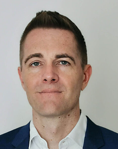 Benjamin Stahl, ein Mann mit kurzen braunen Haaren, trägt ein marineblaues Jackett und ein weißes Hemd. Er steht vor einem schlichten hellen Hintergrund und blickt mit neutralem Gesichtsausdruck direkt in die Kamera.