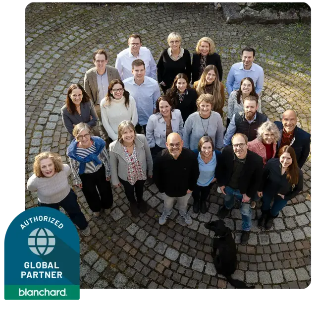 Eine Gruppe von 23 Personen steht im Freien auf einem runden Steinpflaster und lächelt in die Kamera. Ein schwarzer Hund sitzt unter ihnen. In der unteren linken Ecke ist ein Blanchard Global Partner-Abzeichen zu sehen.