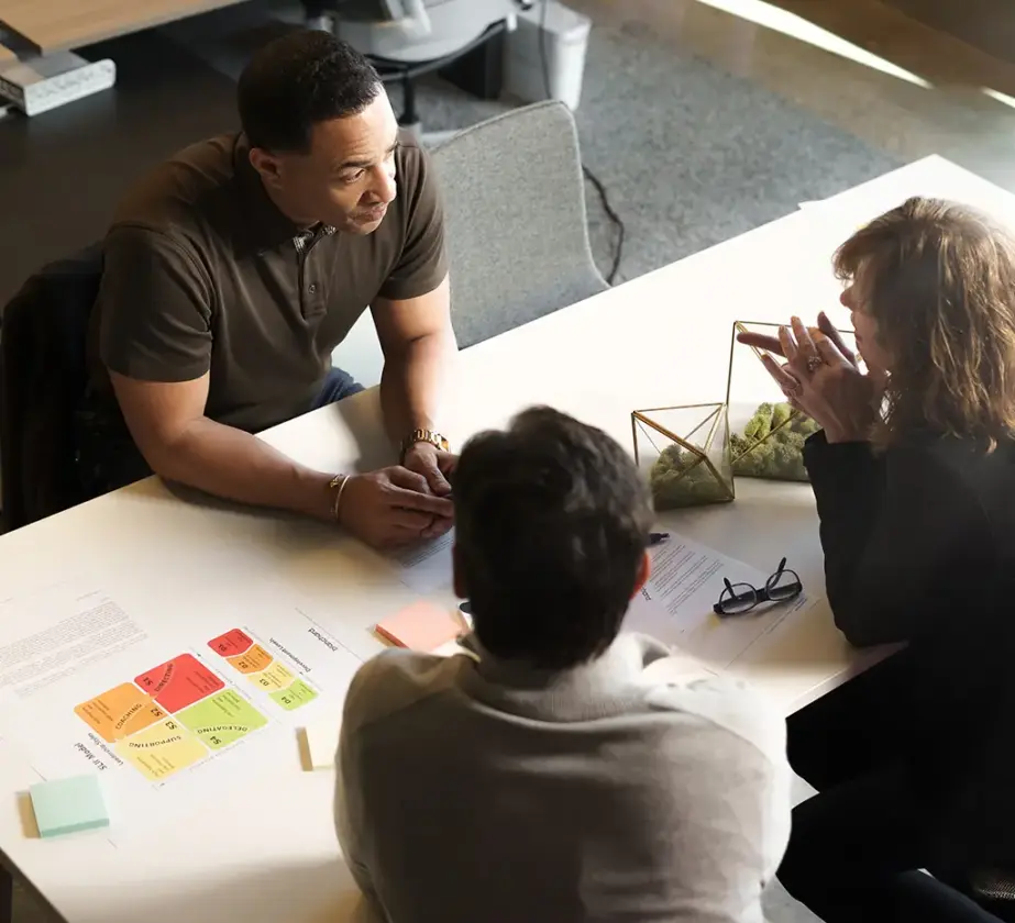 Drei Personen sitzen um einen weißen Tisch und diskutieren. Überall liegen Papiere, bunte Haftnotizen und geometrische Dekorationen herum – eine kollaborative Arbeitsumgebung, die vom Blanchard Führungskräftetraining inspiriert sein könnte.