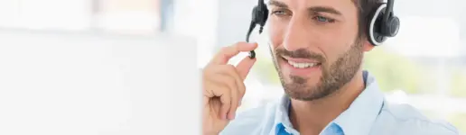 Ein Mann mit Headset und Mikrofon sitzt lächelnd an einem Schreibtisch und blickt auf einen Computerbildschirm. Es scheint, als würde er während eines Online-Meetings Kundensupport leisten oder Cross-Selling betreiben.