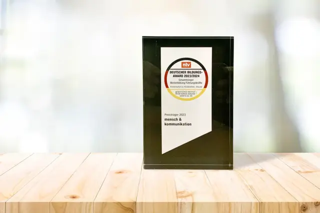 Auf einer hellen Holzoberfläche liegt eine gläserne Auszeichnung mit schwarzem Rand. Der Preis trägt ein kreisförmiges Logo und einen deutschen Text, darunter Deutscher Bildungspreis 2023/2024 und Preisträger 2023 mensch & kommunikation.