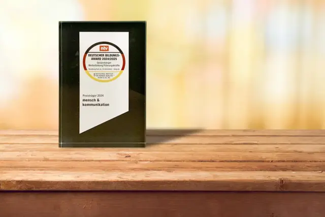 Auf einem Holztisch steht eine gläserne Preisplakette mit der Aufschrift „Deutscher Bildungspreis 2022/2023“, vor einem verschwommenen, warmen Hintergrund. Der Text auf der Plakette beinhaltet Preisträger 2024 mensch & kommunikation.