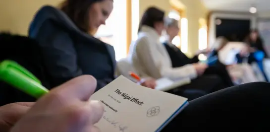 Eine Person schreibt mit einem grünen Stift in ein Notizbuch mit dem Titel „Der Misgi-Effekt“, während andere in einer Reihe neben ihr in einem gut beleuchteten Raum sitzen und sich ebenfalls Notizen machen.