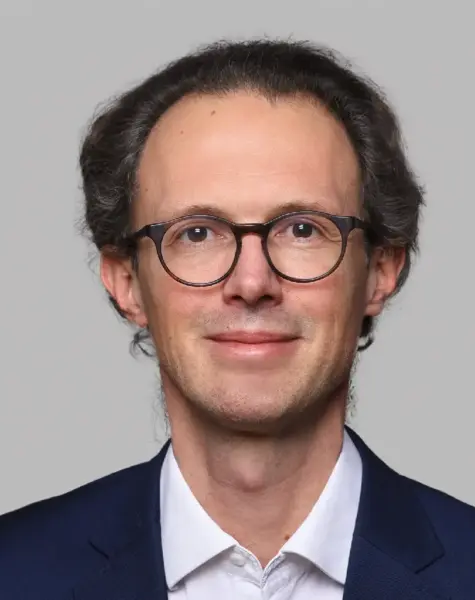 Trainer Fabian Böttger, Experte für Mensch & Kommunikation, trägt einen dunklen Anzug und ein weißes Hemd. Mit kurzem, gut braunem Haar und Brille lächelt er leicht in die Kamera vor einem hellgrauen Hintergrund.