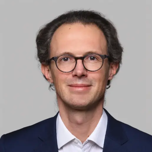 Trainer Fabian Böttger, Experte für Mensch & Kommunikation, trägt einen dunklen Anzug und ein weißes Hemd. Mit kurzem, gut braunem Haar und Brille lächelt er leicht in die Kamera vor einem hellgrauen Hintergrund.