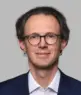 Trainer Fabian Böttger, Experte für Mensch & Kommunikation, trägt einen dunklen Anzug und ein weißes Hemd. Mit kurzem, gut braunem Haar und Brille lächelt er leicht in die Kamera vor einem hellgrauen Hintergrund.