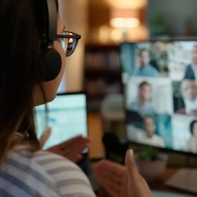 A person wearing headphones and glasses sits at a desk, gesturing while participating in a video conference with several people shown in small windows on a computer screen.