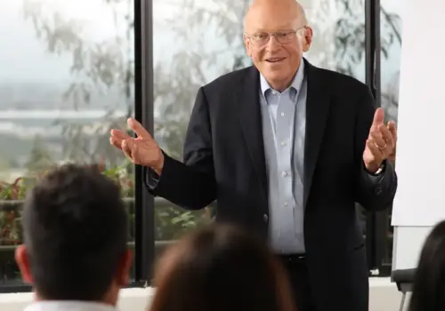 An older man in a suit and glasses speaks to a small group, gesturing with his hands, in front of large windows and a flip chart. The audience faces him, listening attentively.