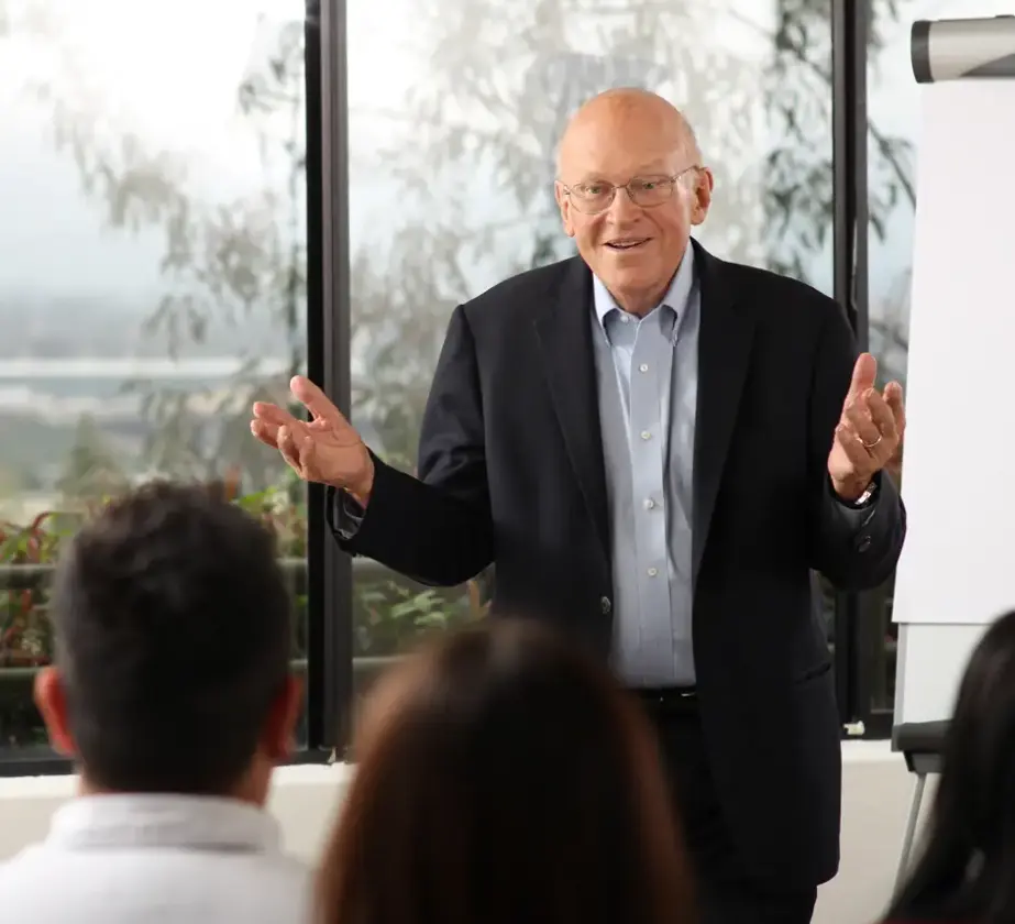 Ken Blanchard steht da und gestikuliert, während er mit einer Gruppe vor ihm sitzender Menschen spricht.
