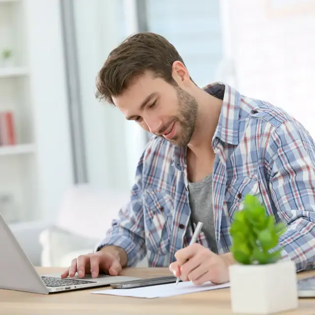 A man in a plaid shirt sits at a desk, using a laptop with one hand and writing on paper with the other. A green potted plant is in the foreground, and the room appears bright and modern.