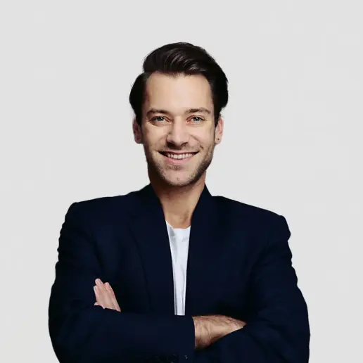 Trainer Manuel Feneberg, Experte für Mensch & Kommunikation, steht lächelnd mit verschränkten Armen im marineblauen Blazer und weißen Hemd vor einem hellgrauen Hintergrund.