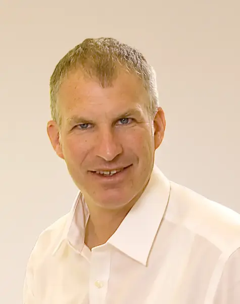 A middle-aged man with short gray hair is smiling and wearing a white collared shirt, posing in front of a plain light-colored background.