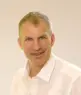 Trainer Martin Lunz, ein Mann mittleren Alters mit kurzen grauen Haaren, lächelt in einem weißen Hemd vor einem schlichten hellen Hintergrund in die Kamera.
