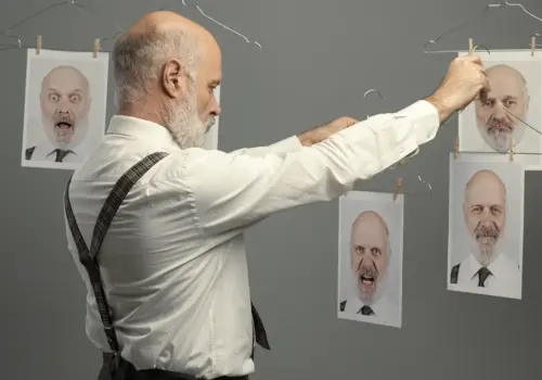 Ein Mann mit Bart und Hosenträgern hängt gedruckte Fotos seines eigenen Gesichts, die jeweils unterschiedliche Emotionen zeigen, an eine Wäscheleine vor einem schlichten grauen Hintergrund und symbolisiert, wie wir unsere emotionale Intelligenz entwickeln können.