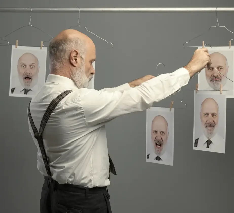 Ein Mann mit Bart und Hosenträgern hängt gedruckte Fotos seines eigenen Gesichts, die jeweils unterschiedliche Emotionen zeigen, an eine Wäscheleine vor einem schlichten grauen Hintergrund und symbolisiert, wie wir unsere emotionale Intelligenz entwickeln können.