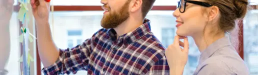 Ein Mann und eine Frau stehen an einem Whiteboard und erzählen gemeinsam Geschichten. Der Mann schreibt mit einem Marker, während die Frau mit Brille nachdenklich zusieht. Beide lächeln und scheinen in einem hellen Büro in eine Diskussion vertieft zu sein.